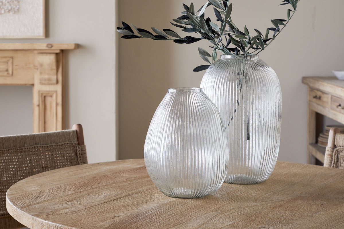 Two Linsy Ribbed Glass Vases by nkuku rest on a round wooden table. The larger clear vase displays olive branches, while the smaller is empty. Light wood furniture and a neutral wall add to the calm, rustic atmosphere.