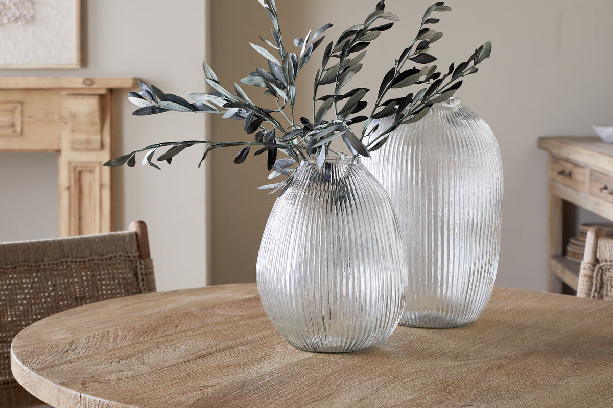 Two Linsy Ribbed Glass Vases by nkuku sit on a round wooden table. One clear vase holds green leafy branches, with wooden furniture and neutral walls in the background for a calm, rustic look.
