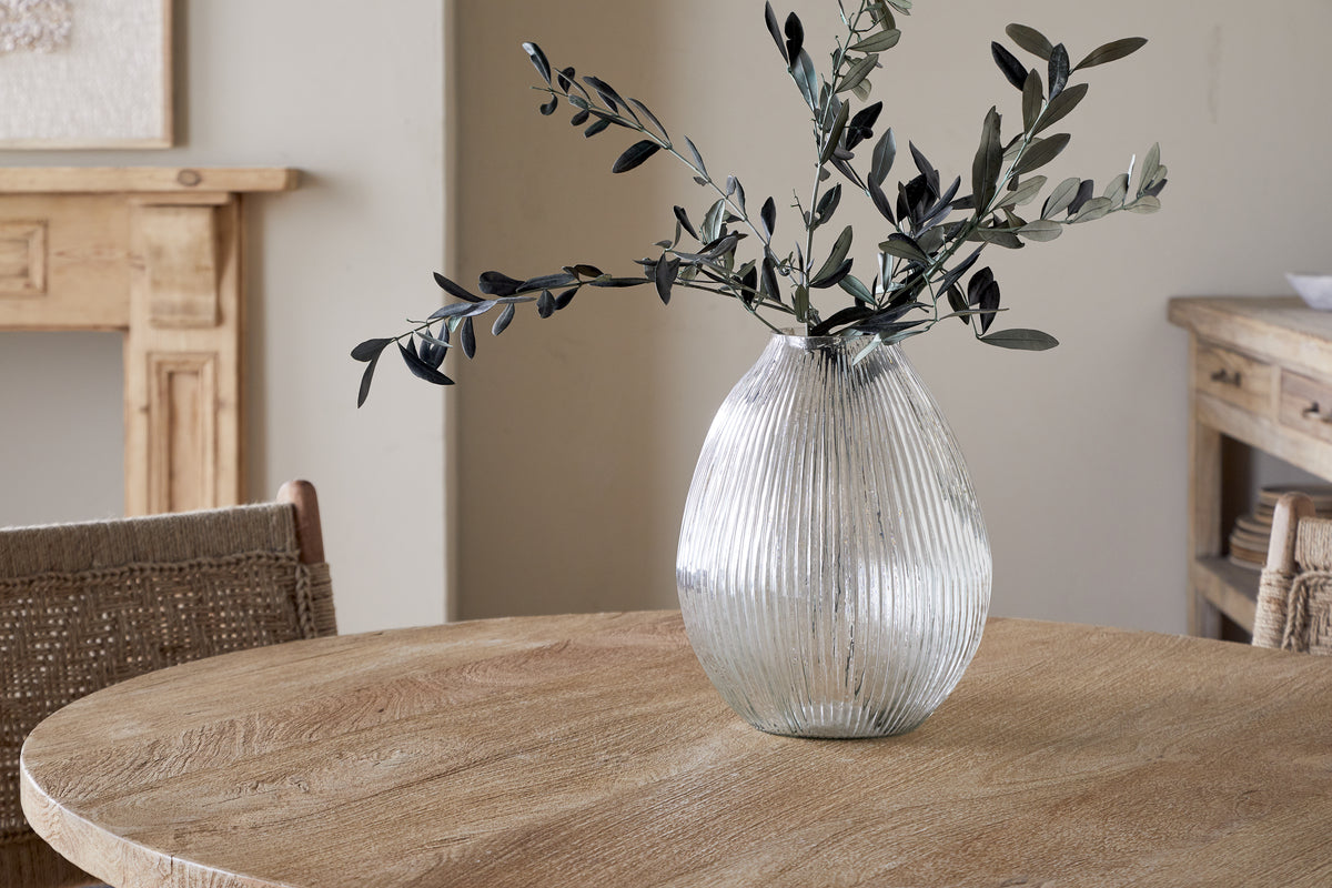 The nkuku Linsy Ribbed Glass Vase - Clear with olive branches stands on a round wooden table. Woven chairs and a light wood sideboard enhance the cozy, minimalist room with neutral walls.