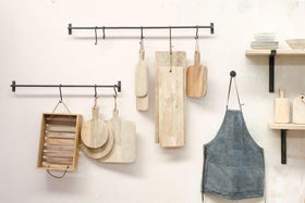The nkuku Laila Hanging Rail in iron holds wooden cutting boards, a washboard, and an apron on hooks. A nearby wooden shelf with stacked white dishes adds to the organized, stylish look against the light wall in your kitchen.