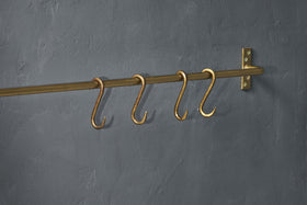 Four Laila Iron S-Hooks in a brass finish by nkuku are evenly spaced on a traditional brass rod, mounted to a textured dark gray wall with a matching right-side bracket; no items hang from the S-hooks.