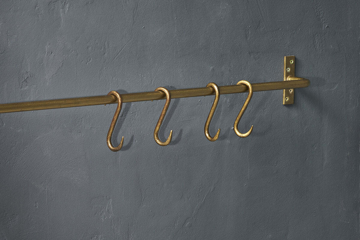 Four Laila Iron S-Hooks in a brass finish by nkuku are evenly spaced on a traditional brass rod, mounted to a textured dark gray wall with a matching right-side bracket; no items hang from the S-hooks.