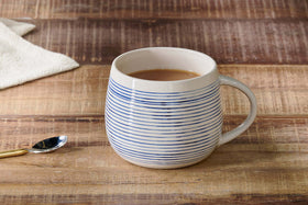 The Lin Oversized Mug by nkuku, featuring blue and white stripes, sits on a wooden surface filled with coffee or tea. Nearby rests a metal spoon with a gold handle and a folded cream-colored napkin—ideal for cosy drinks.