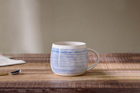 The Lin Oversized Mug by nkuku, featuring horizontal blue stripes and a right-side handle, sits on a wooden table—perfect for cozy drinks. A silver spoon and folded white napkin are softly blurred in the background.