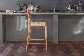 The Lohanda Acacia & Munja Grass Counter Chair by nkuku stands before a gray concrete island in a modern, minimalist kitchen with potted plants, wooden boards, jars, and tableware on dark herringbone wood floors.