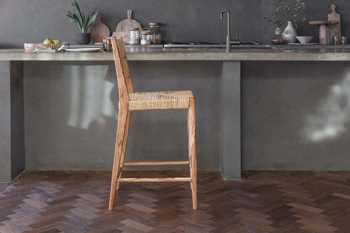 The Lohanda Acacia & Munja Grass Counter Chair by nkuku stands before a gray concrete island in a modern, minimalist kitchen with potted plants, wooden boards, jars, and tableware on dark herringbone wood floors.
