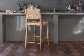 The nkuku Lohanda Acacia & Munja Grass Counter Chair - Natural stands before a gray concrete kitchen counter topped with plates, bowls, chopping boards, and a small plant above dark herringbone wood flooring.