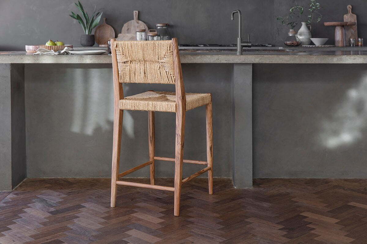 The nkuku Lohanda Acacia & Munja Grass Counter Chair - Natural stands before a gray concrete kitchen counter topped with plates, bowls, chopping boards, and a small plant above dark herringbone wood flooring.
