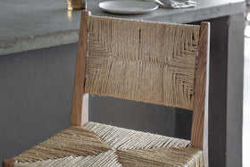 Close-up of the nkuku Lohanda Acacia & Munja Grass Counter Chair in natural, featuring a wooden frame and woven seat, next to a grey concrete dining table set with a white plate and cutlery.