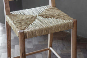 The nkuku Lohanda Acacia & Munja Grass Counter Chair features a handcrafted acacia frame and a seat intricately woven from natural beige munja grass, highlighting artisanal craftsmanship and elegant geometric patterns.