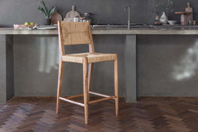 The Lohanda Acacia & Munja Grass Counter Chair by nkuku, featuring a woven seat and back, sits on herringbone parquet before a concrete kitchen island with a sink, cutting boards, and plants in the background.