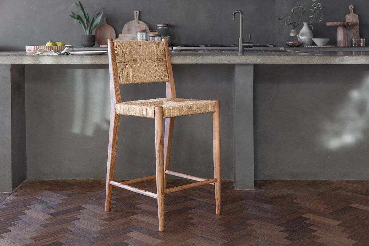 The Lohanda Acacia & Munja Grass Counter Chair by nkuku, featuring a woven seat and back, sits on herringbone parquet before a concrete kitchen island with a sink, cutting boards, and plants in the background.