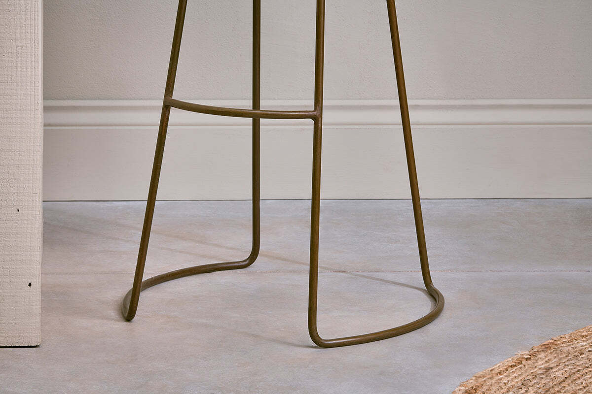 Close-up of the Loko Mango Wood Stool - Tall by nkuku, featuring a modern rustic iron base with thin curved legs and a footrest on a smooth light gray floor, against a white wall with trim and part of a woven rug in the corner.