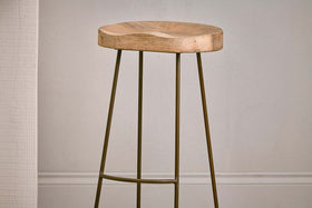 The Loko Mango Wood Stool - Tall by nkuku features a minimalist design with a rounded, smooth mango wood seat and three slender outward-angled metal legs, set against a plain light wall with white trim.