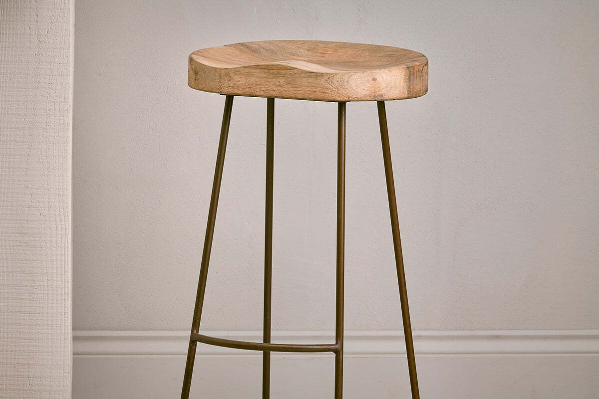 The Loko Mango Wood Stool - Tall by nkuku features a minimalist design with a rounded, smooth mango wood seat and three slender outward-angled metal legs, set against a plain light wall with white trim.