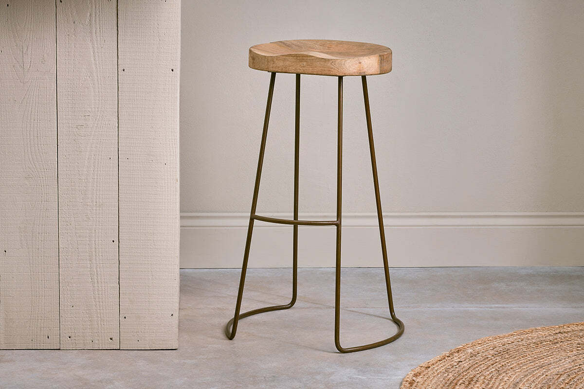 The nkuku Loko Mango Wood Stool - Tall features a round, carved mango wood seat and slim dark metal legs, set on a grey concrete floor beside a light vertical wooden panel.