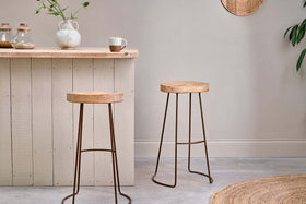 Two Loko Mango Wood Stools by nkuku, with dark iron legs and sustainable mango wood seats, stand in front of a light wood bar. A textured white vase with greenery and a two-tone mug complete the inviting scene.