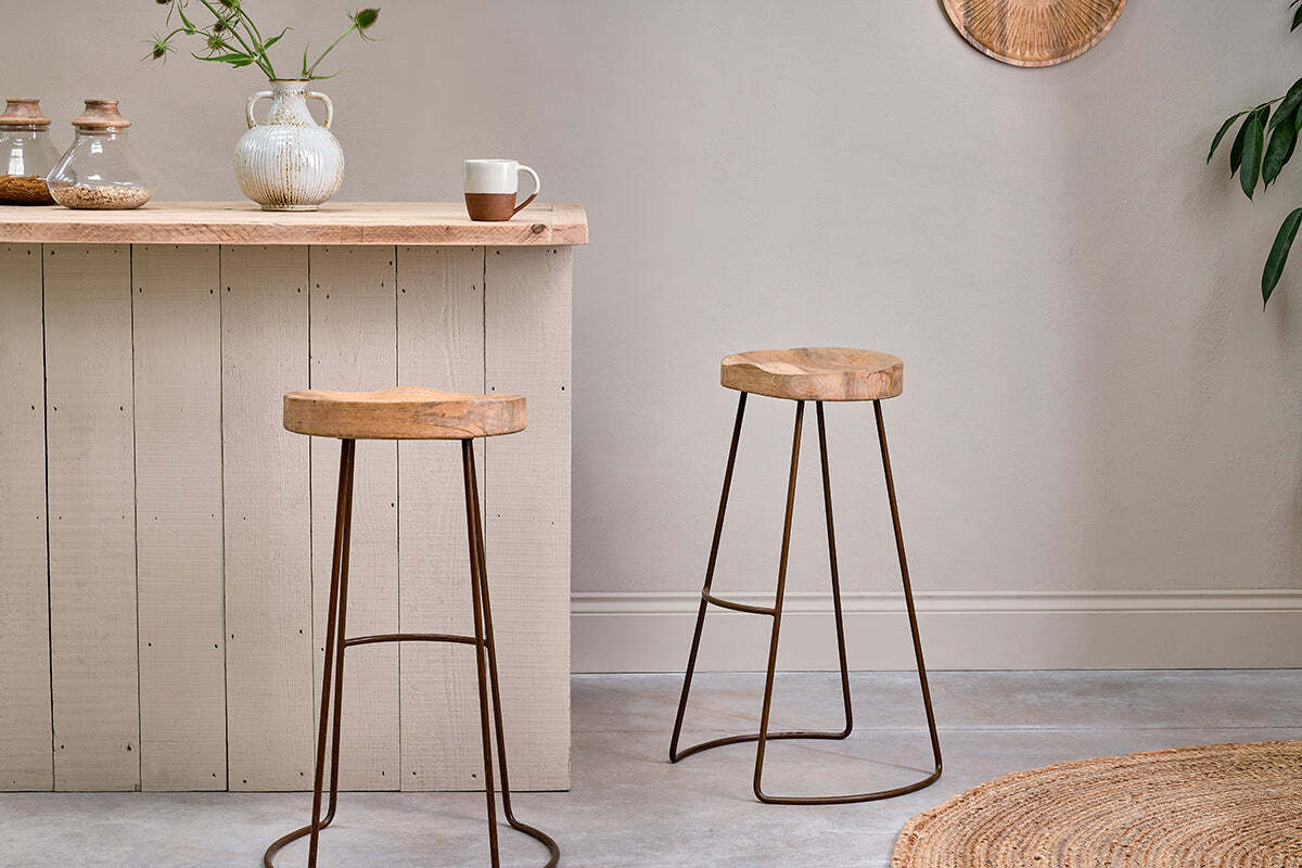 A minimalist kitchen showcases a light wooden counter with jars, a vase, and a mug. Two nkuku Loko Mango Wood Stools - Tall add rustic charm with iron frames and wooden seats, while a woven rug and potted plant accent the neutral decor.