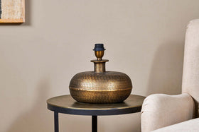 The Lumbu Lamp - Antique Brass - Small by nkuku, with its metallic textured artisan base, sits perfectly on a side table next to a beige chair, adding stylish decorative lighting to any space.
