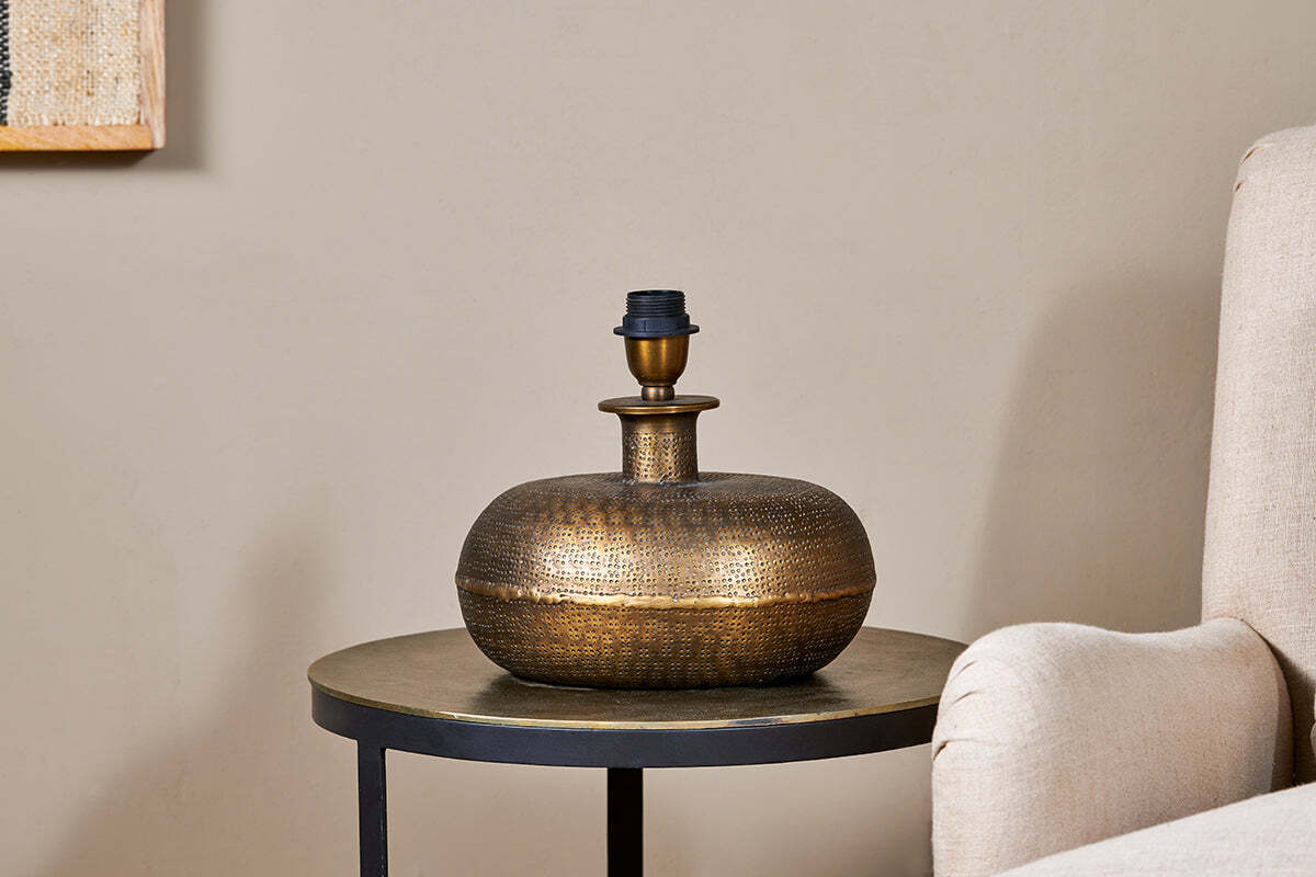 The Lumbu Lamp - Antique Brass - Small by nkuku, with its metallic textured artisan base, sits perfectly on a side table next to a beige chair, adding stylish decorative lighting to any space.