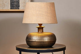 The nkuku Lumbu Lamp in Antique Brass (Small) with a wide, textured base and beige fabric shade sits on a round, dark side table against a neutral wall. Turned on, it casts warm decorative light. A picture frame is visible in the background.