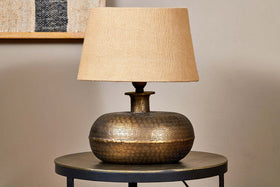 The nkuku Lumbu Lamp in antique brass, small size, with a textured base and beige fabric shade sits on a metallic side table. A neutral wall and framed artwork in the background highlight this elegant decorative lighting piece.