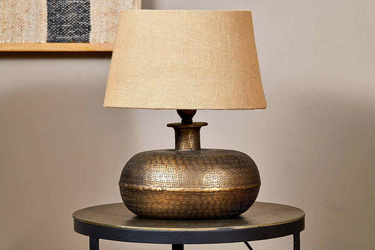 The nkuku Lumbu Lamp in antique brass, small size, with a textured base and beige fabric shade sits on a metallic side table. A neutral wall and framed artwork in the background highlight this elegant decorative lighting piece.