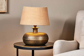 The nkuku Lumbu Lamp in Antique Brass (Small), featuring a textured base and beige shade, sits on a black side table by a light beige armchair against a neutral wall. Its warm glow adds soft decorative lighting to the space.