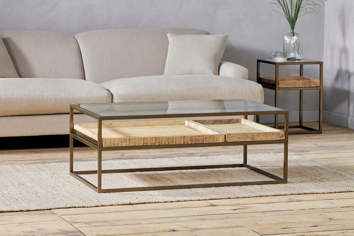 The nkuku Luzon Iron & Mango Wood Display Coffee Table – Brass sits on a woven rug in front of a sectional sofa, with a side table featuring wooden shelves in the background.