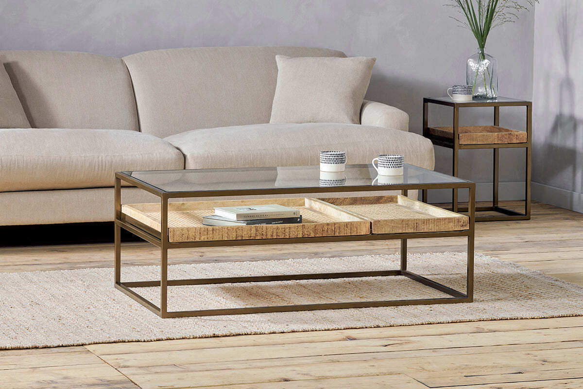 A modern living room featuring the nkuku Luzon Iron & Mango Wood Display Coffee Table in brass, topped with mango wood trays, striped cups, and books. A small side table holds a glass vase and plant over a light rug on wooden floors.