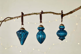 Three indigo glass Manipur Baubles by nkuku, featuring ribbed textures and brass tops, hang from dark ribbons. The set of three ornaments—diamond, teardrop, and round—adds vintage charm to your festive decor.