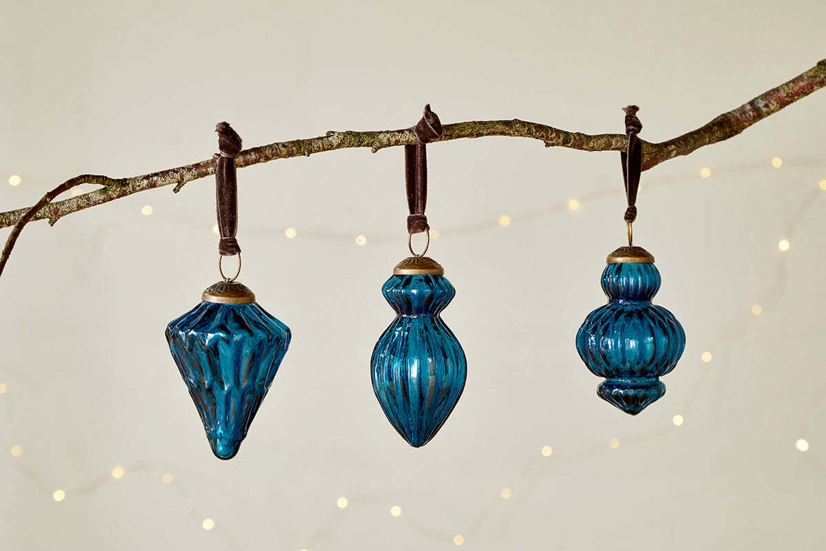 Three indigo glass Manipur Baubles by nkuku, featuring ribbed textures and brass tops, hang from dark ribbons. The set of three ornaments—diamond, teardrop, and round—adds vintage charm to your festive decor.