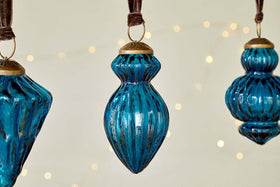 The Manipur Baubles - Indigo (Set of 3) by nkuku feature ribbed blue glass with gold tops, hanging from brown ribbons. Their vintage-inspired shapes and warm white fairy lights in the background create a cozy festive décor.
