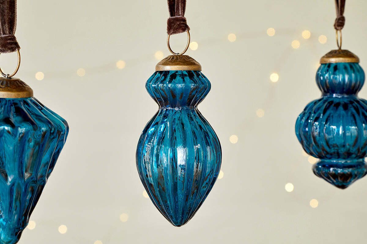 The Manipur Baubles - Indigo (Set of 3) by nkuku feature ribbed blue glass with gold tops, hanging from brown ribbons. Their vintage-inspired shapes and warm white fairy lights in the background create a cozy festive décor.