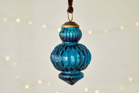 The nkuku Manipur Baubles - Indigo (Set of 3) feature blue ribbed glass with brass caps and loops, hanging from brown cords. Their vintage style and warm glow evoke classic festive charm against a cream background.