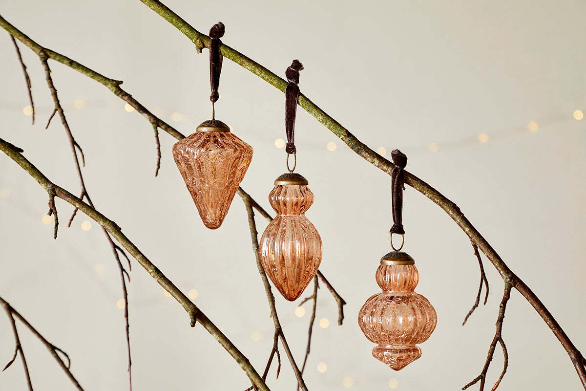 Three ribbed copper Manipur Baubles from nkuku, each with a bronze top and brown ribbon loop, hang on bare branches. Vintage Christmas decor and blurred warm fairy lights in the background create a festive, elegant scene.