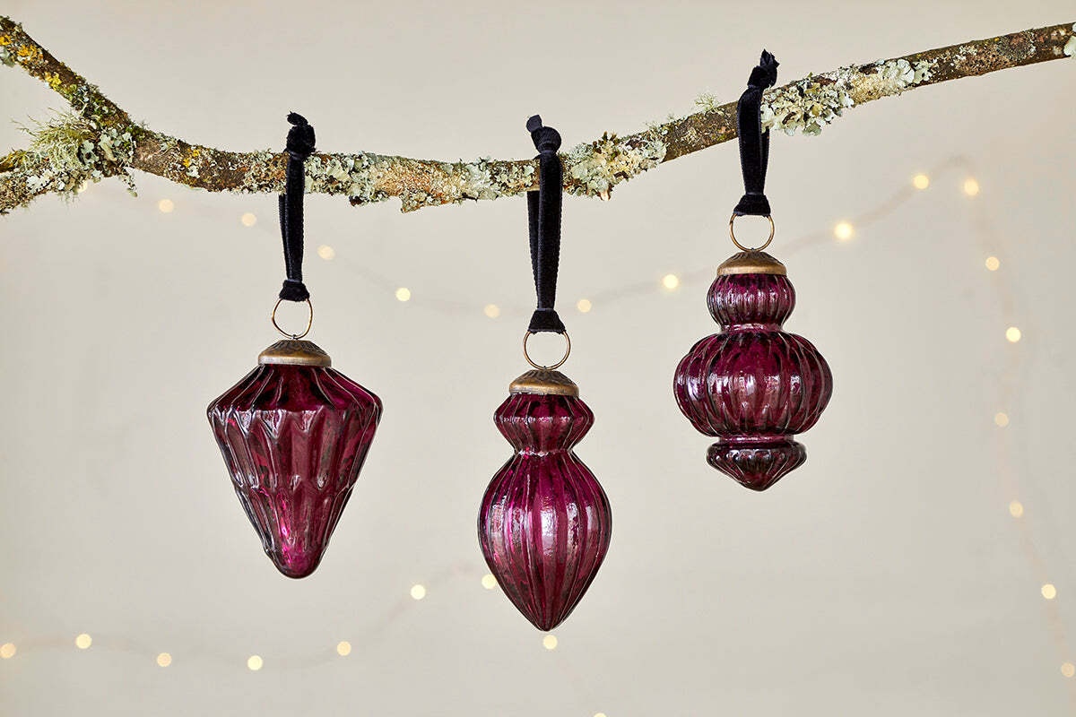 A set of three deep ruby Manipur Baubles by nkuku hangs from black ribbons on a lichen-covered branch. Gold caps and a soft beige background with blurred fairy lights create a cozy, festive atmosphere.