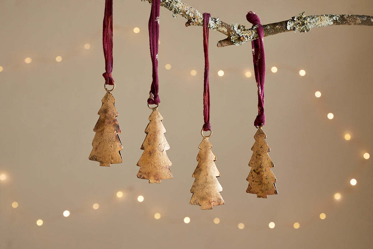 The Mishti Tree Baubles - Antique Brass - Small (Set of 4) by nkuku hang from a lichen-covered branch on burgundy ribbons, with a warm, softly blurred background and glowing lights for a festive, cozy feel.