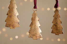Four Mishti Tree Baubles in antique brass by nkuku hang on maroon ribbons against a soft, warm backdrop with blurred twinkling lights, adding a festive and cozy touch to your holiday decor.