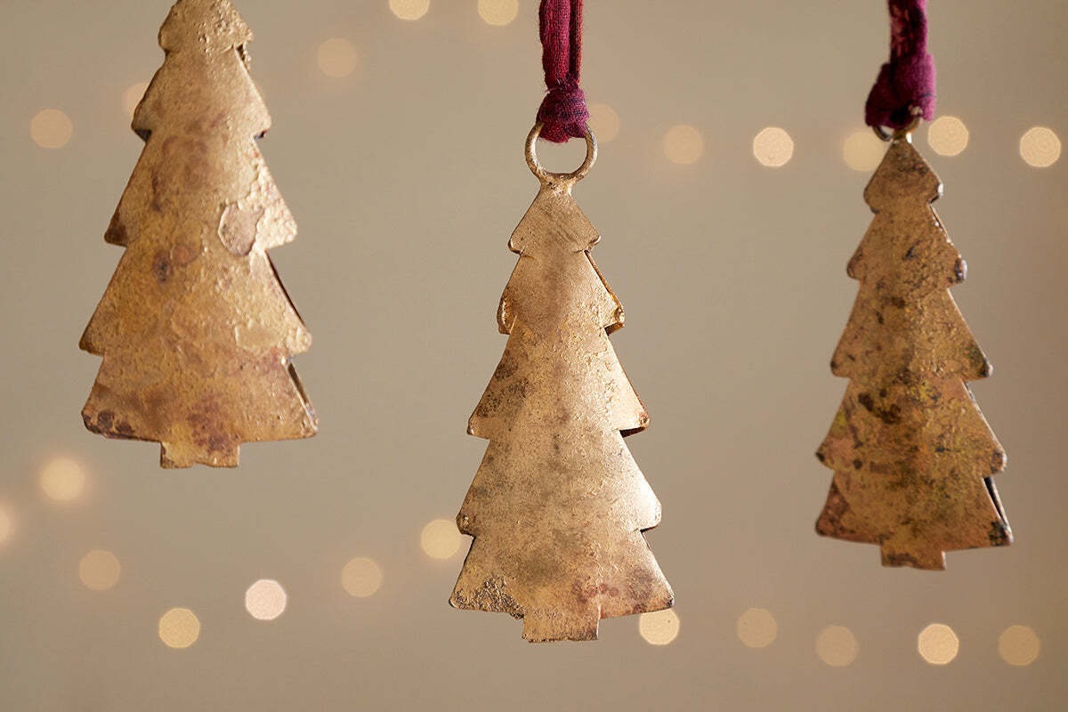 Four Mishti Tree Baubles in antique brass by nkuku hang on maroon ribbons against a soft, warm backdrop with blurred twinkling lights, adding a festive and cozy touch to your holiday decor.