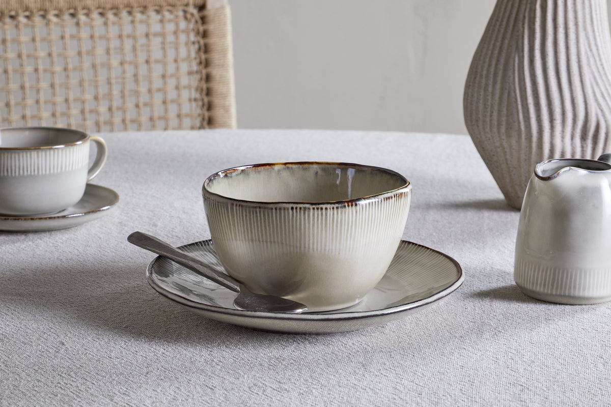 The Malia Breakfast Bowl in Cream by nkuku sits atop a white tablecloth, its organic shape mirrored by the woven-back chair nearby, while coordinated ceramics and a textured vase add subtle charm to the scene.