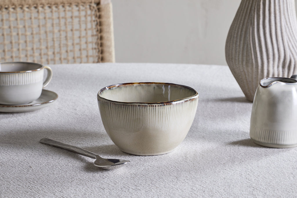 The nkuku Malia Breakfast Bowl - Cream, featuring a reactive glaze and organic shape, rests on a textured white tablecloth alongside a metal spoon, cup and saucer, small pitcher, ribbed vase, and a woven chair in the background.