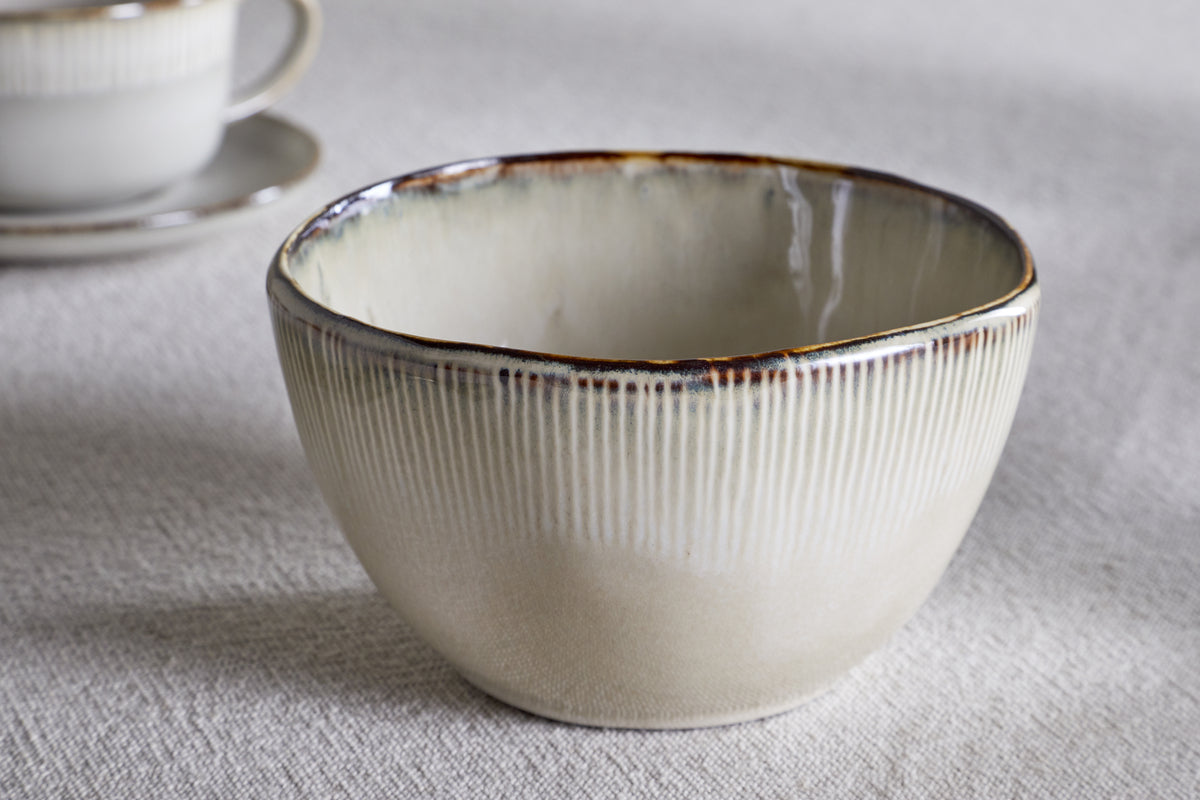 A cream Malia Breakfast Bowl by nkuku, featuring a gently irregular shape and vertical ridges, sits on a light textured surface. In the background, a matching cup and saucer with a subtle reactive glaze are softly blurred.