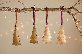 A lichen-covered branch displays four nkuku Mishti Tree Baubles in antique brass, each large bauble hung by maroon or orange ribbon. Warm white fairy lights blur softly behind, creating a charming festive scene.