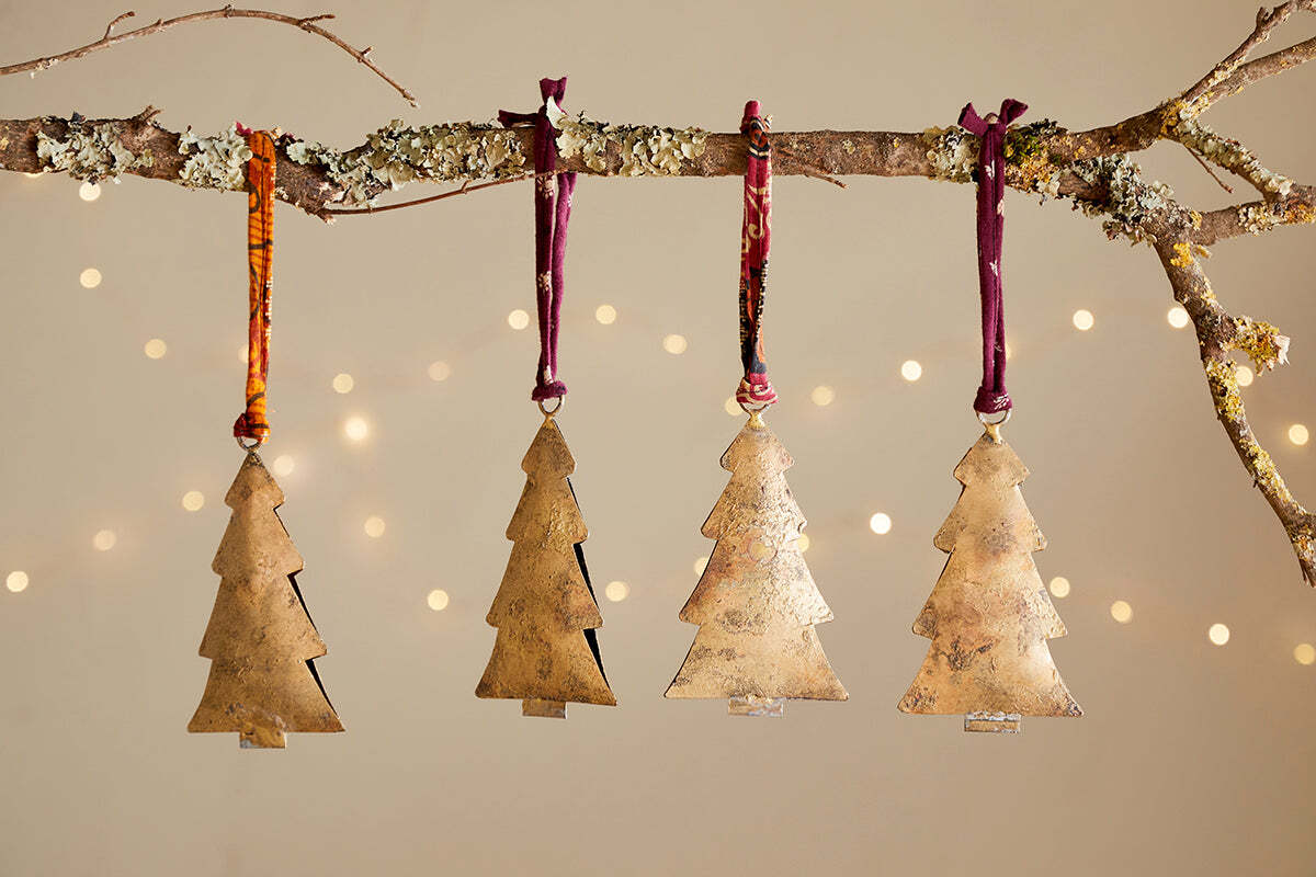 A lichen-covered branch displays four nkuku Mishti Tree Baubles in antique brass, each large bauble hung by maroon or orange ribbon. Warm white fairy lights blur softly behind, creating a charming festive scene.