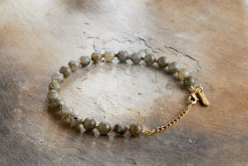 The Marai Bracelet - Labradorite by nkuku features round, mottled greenish-grey labradorite stones and a gold chain clasp, elegantly presented on a textured wooden surface.
