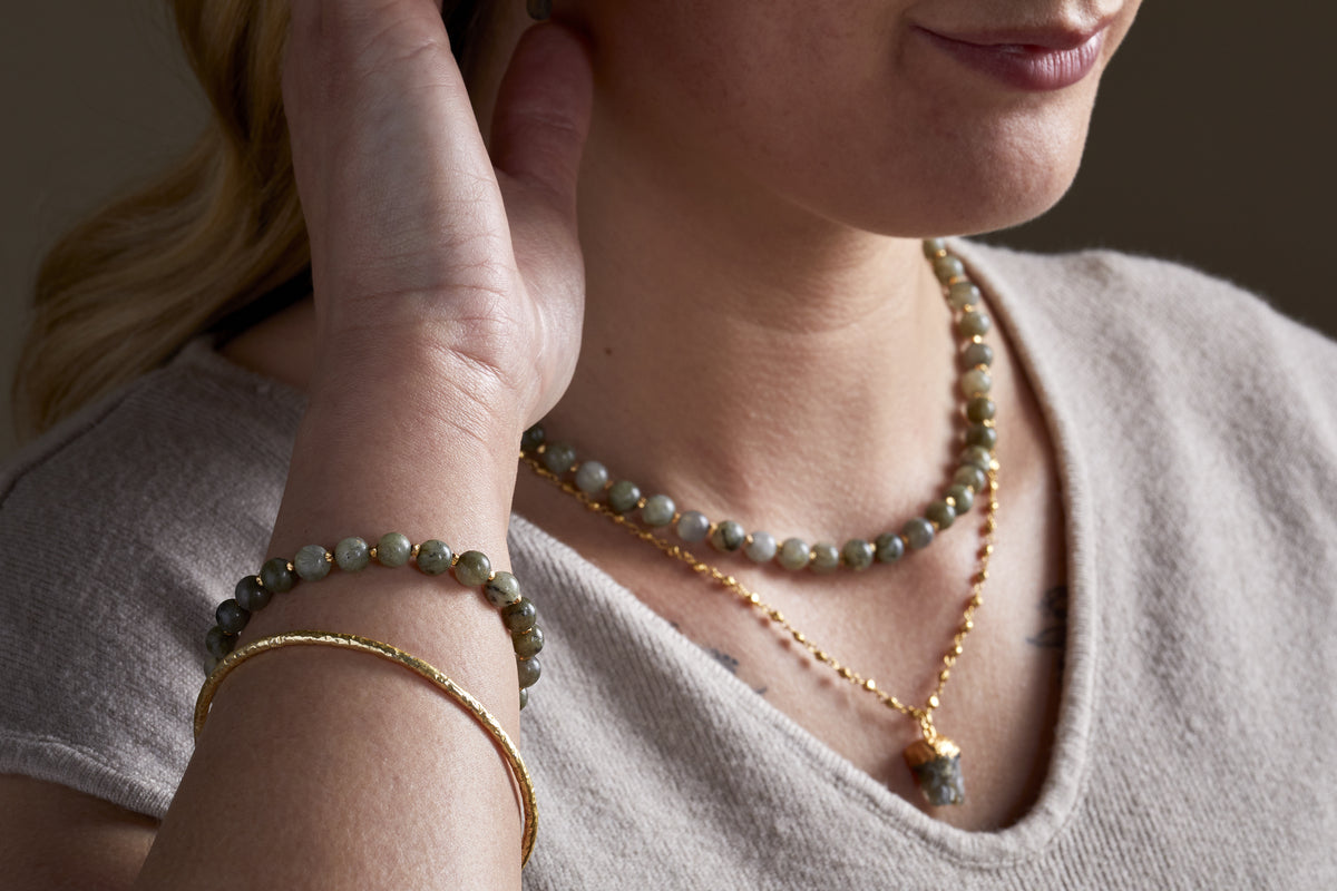A woman wearing a light beige top and elegant jewellery, including nkuku’s Marai Bracelet – Labradorite, touches her ear with one hand.