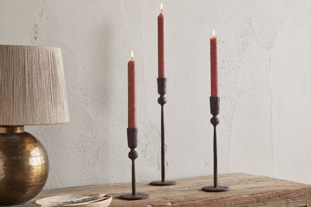 A wooden table features three lit red taper candles in nkuku Mawindi Candle Holders - Rust beside a round bronze lamp with a beige shade, set against a textured light wall for an earthy decor accent.