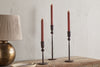 Three red taper candles in nkuku’s Mawindi Candle Holders - Rust are displayed at different heights on a rustic wood surface beside a beige and brass lamp, all set against a textured light wall for an earthy decor accent.