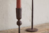 The Mawindi Candle Holder - Rust by nkuku is shown with a red candle on a wooden surface against a textured wall, with another blurred holder in the background for an earthy touch.